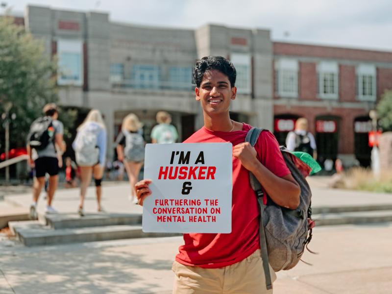 Naren smiles for a photo holding a sign that says "I'm a Husker & furthering the conversation on mental health"