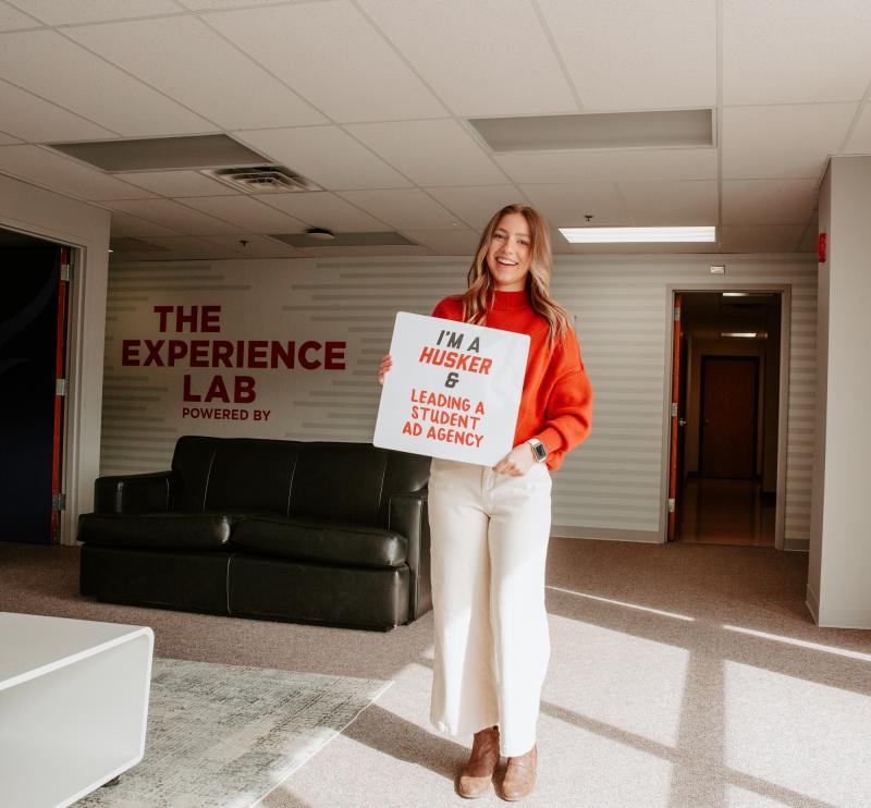 Paige smiles for a photo in the Experience Lab holding a sign that says “I’m a Husker & leading a student ad agency”