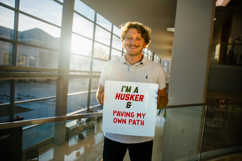 Samuel smiles for photo with a sign that says “I’m a Husker & paving my own path”
