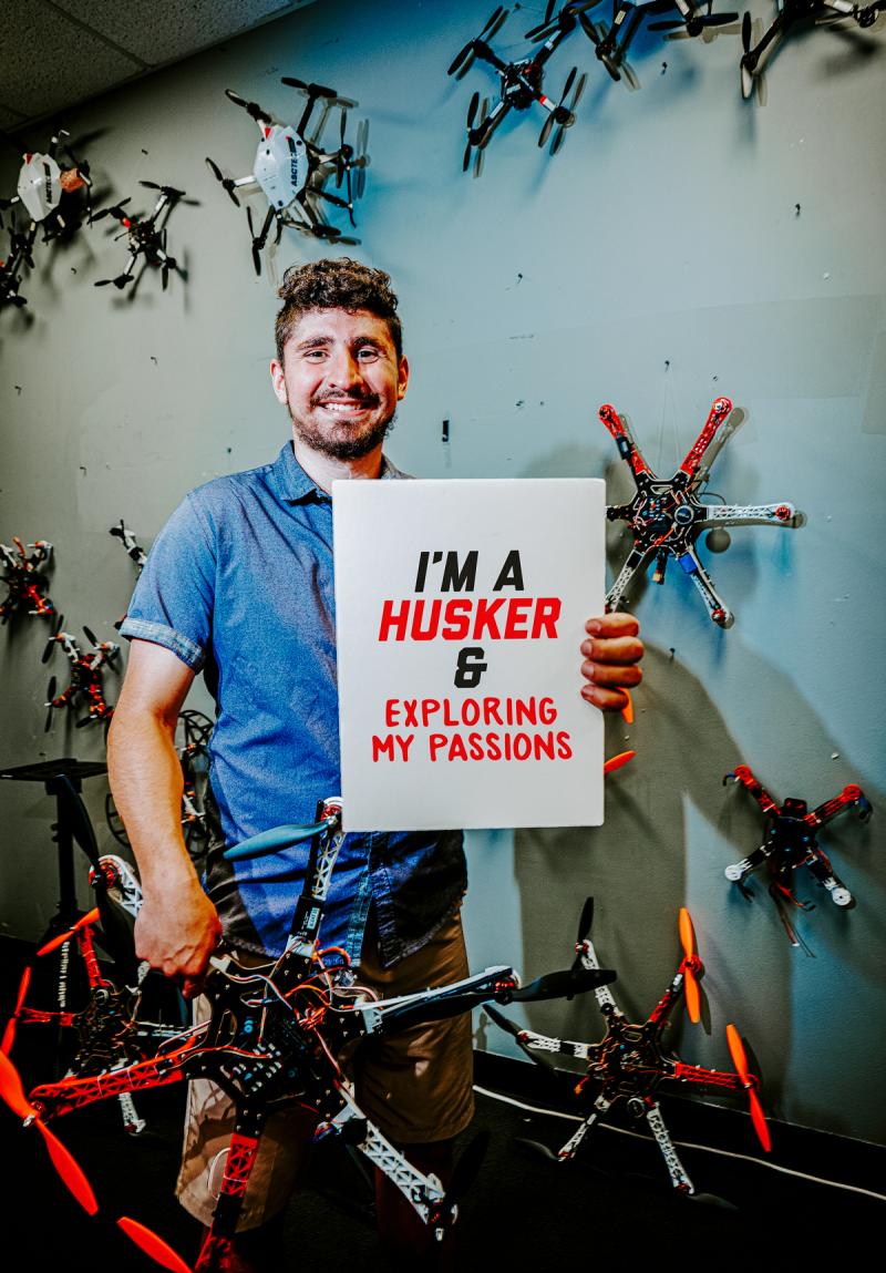 Santiago stands in the NIMBUS lab surrounded by drones and holding a drone in his right hand. In his left hand he holds a sign that reads "I'm a Husker and exploring my passions"