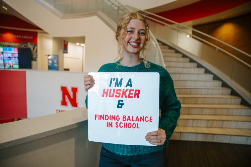 Saylor smiles for a photo in the Union holding a sign that says “I’m a Husker & finding balance in school”