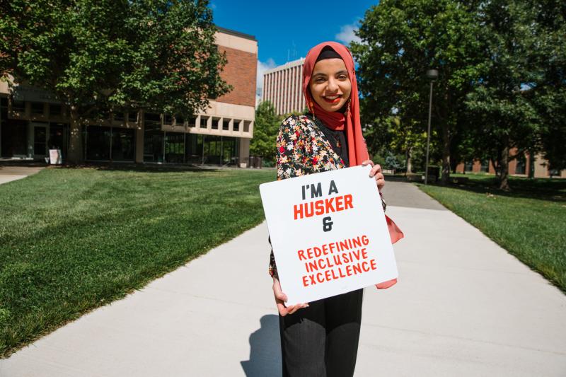 Sukaina smiles for a photo with a sign that says “I’m a Husker & redefining inclusive excellence”