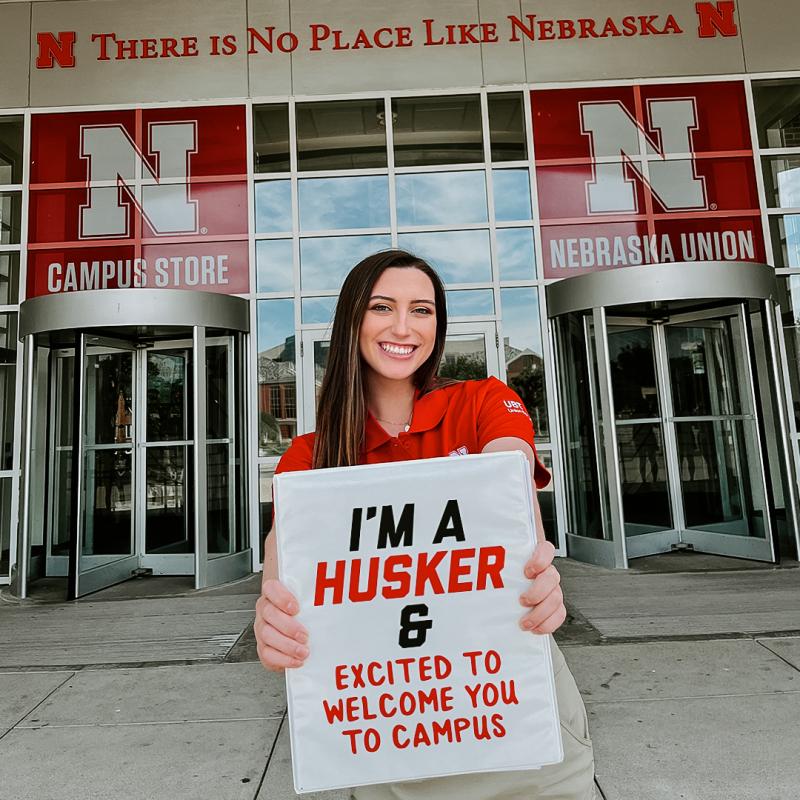 Sydney stands in front of the Nebraska Union holding a sign that reads, "I'm a Husker, and excited to welcome you to campus!"