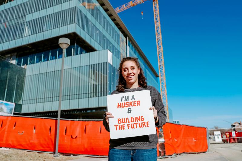 Taylor smiles for a photo outside the new College of Engineering building being built. She holds a sign that reads “I’m a Husker & building the future”