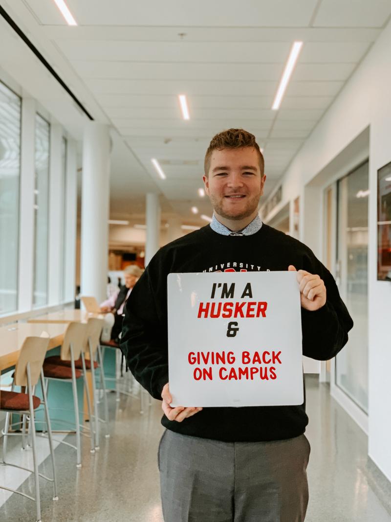 Troy smiles for a photo in the Health Center with a board that says "I'ma Husker & giving back on campus"