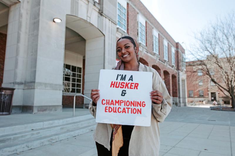 Yousra smiles for a photo outside the Union with a board that reads "I'm a Husker & championing education"