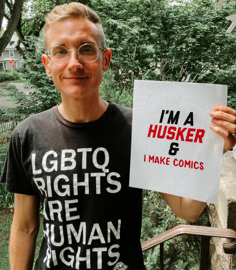 James holds up a sign that says "I'm a Husker & I make comics"