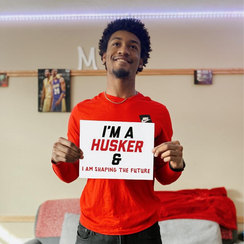 Michael smiles in his room holding a sign that says “I’m a Husker & I am shaping the future”