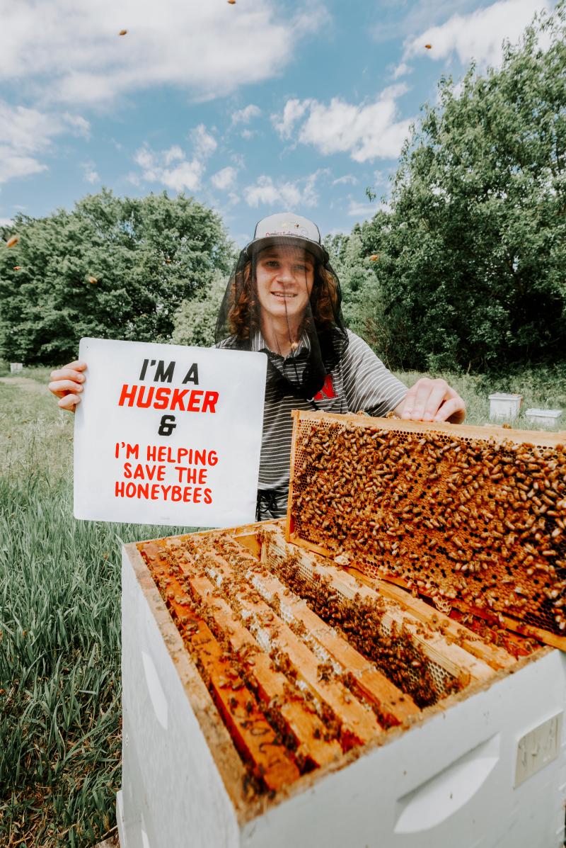 Rogan Rokach stands with a bee hive and a bee net holding a sign that reads "I'm a Husker and I'm helping save the honey bees!"