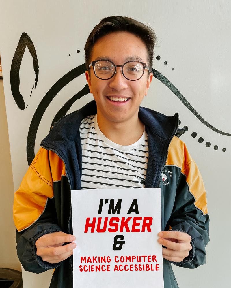 Keith smiles for a photo holding a sign that says "I'm a Husker & making computer science accessible."