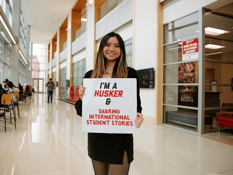 Jermaine smiles for a photo in Hawks Hall holding a sign that says “I’m a Husker & sharing international student stories”
