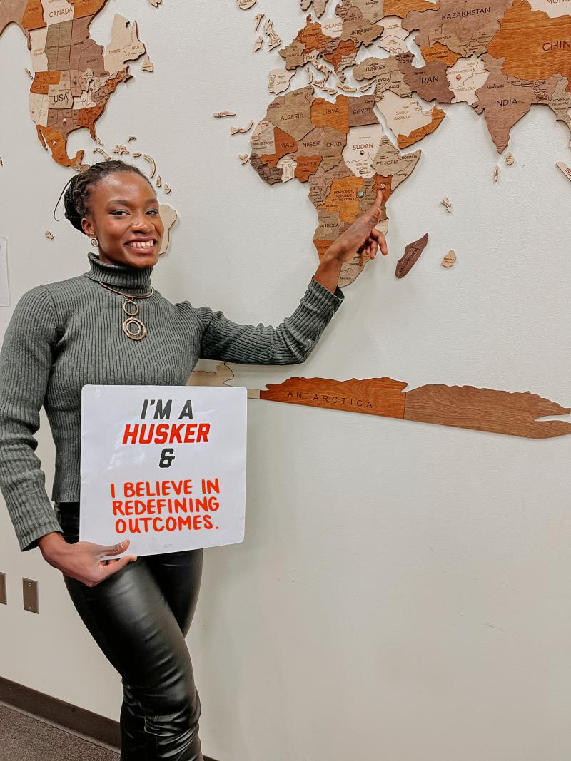 Rosemary Onyango points to her home country, Kenya, on the map. She holds a sign reading "I'm a Husker and I believe in redefining outcomes."